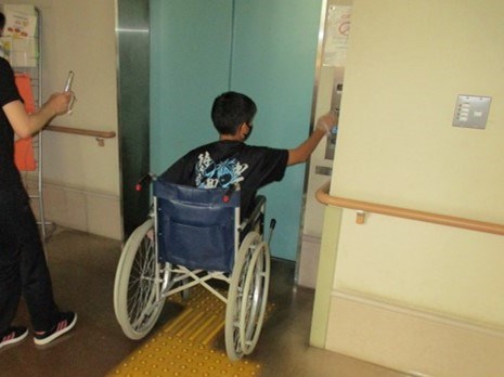 車いす体験の画像です。車いすに乗ったまま子どもがエレベータのスイッチを押そうとしています。